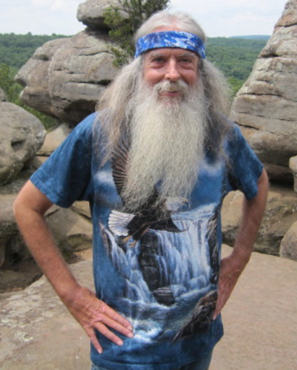 Picture of Bill Choisser standing on a rocky overlook with a forest of trees in the background. He is smiling with his hands on his hips and is wearing a blue t-shirt with a bald eagle flying in front of a waterfall and a matching blue tie dye bandana across his forehead, tying back long white hair. He has a matching long white beard that goes down to the middle of his chest.